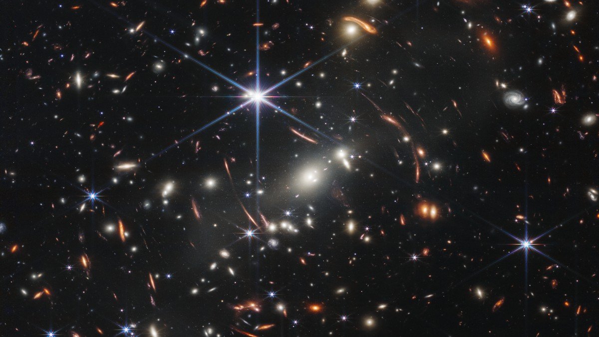 Deep space astronomical image showing a compact reddish elliptical galaxy surrounded by faint distant cosmic structures